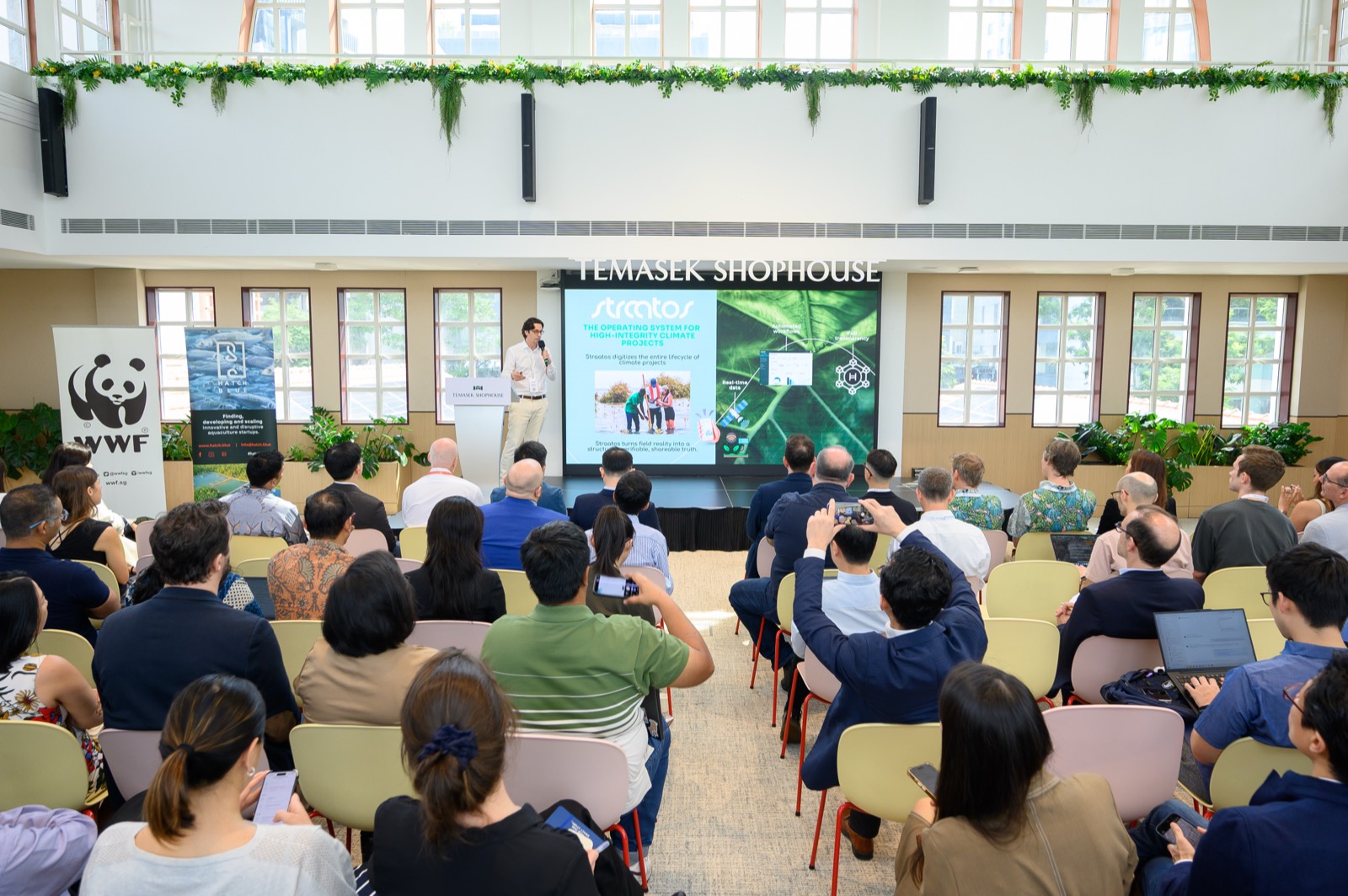 A Straatos presenter on stage at the Blue Catalyst Challenge Demo Day, with a slide reading "The operating system for high-integrity climate projects", before a seated audience at Temasek Shophouse.